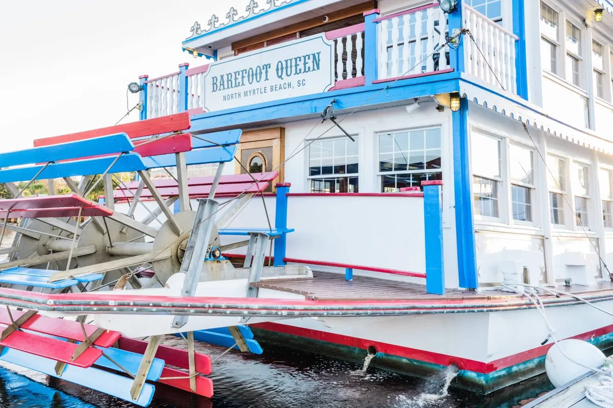 This exterior image features the colorful paddle wheel of the Barefoot Queen riverboat, docked along the scenic waterfront. The lively boat is shown with its signature signage and vibrant colors, inviting guests aboard for sightseeing, dining, and socializing. The setting sun casts a warm glow on the water, enhancing the sense of tranquil luxury and unique local experiences.