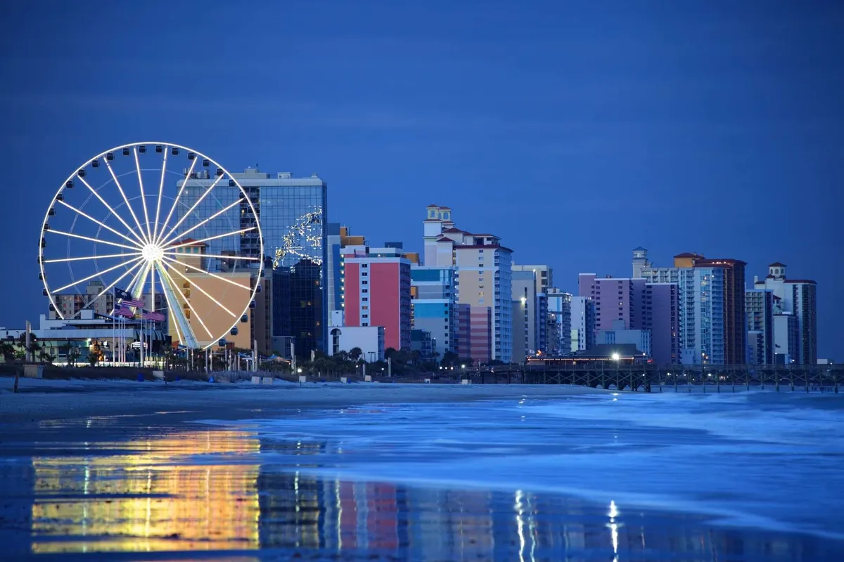 The image features a vibrant Ferris wheel set against a backdrop of colorful buildings, with reflections glimmering on the water below. The scene is lively, evoking a classic beach boardwalk atmosphere, and hints at seaside fun with rides, nearby restaurants, and the ocean just beyond.