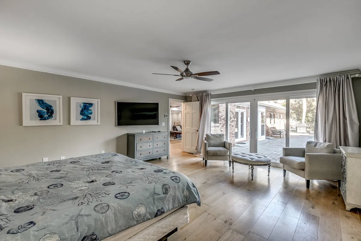 A spacious bedroom featuring a comfortable bed with patterned duvet, an armchair, a television, sliding glass doors leading outside, and artwork on the wall. The room is flooded with natural light, creating an inviting and calm atmosphere ideal for relaxation.