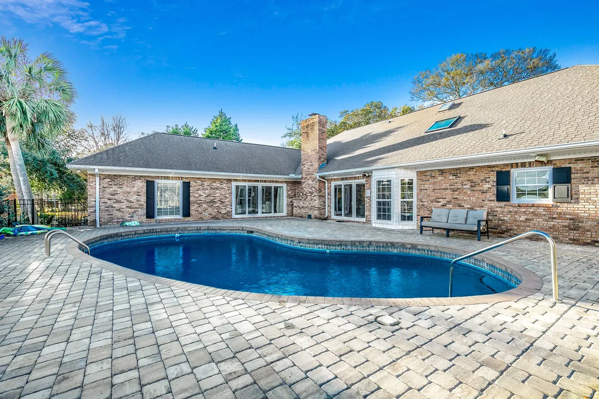 A sunlit backyard scene featuring a sparkling swimming pool surrounded by a brick house exterior. The patio area is furnished with ample outdoor seating, suggesting a space for sunbathing, swimming, and relaxed family gatherings. The pool area is lively, with children playing in the water and adults lounging nearby, evoking an inviting, resort-like ambiance.