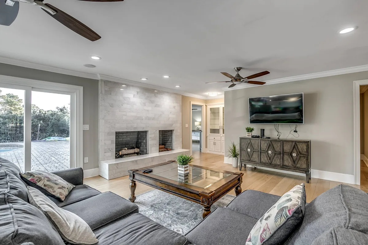 A spacious living room featuring a plush sofa with decorative pillows, a glass coffee table, dual fireplaces, a flatscreen television mounted on the wall, and sliding doors that open to the outside. The space is bright and airy, feeling both cozy and expansive—perfect for gatherings and evening relaxation.