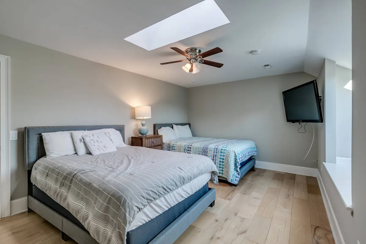 A cozy bedroom with two beds under a skylight, a ceiling fan, and a nightstand with a lamp. The room exudes comfort and is ideal for flexible sleeping arrangements, such as for families or groups.