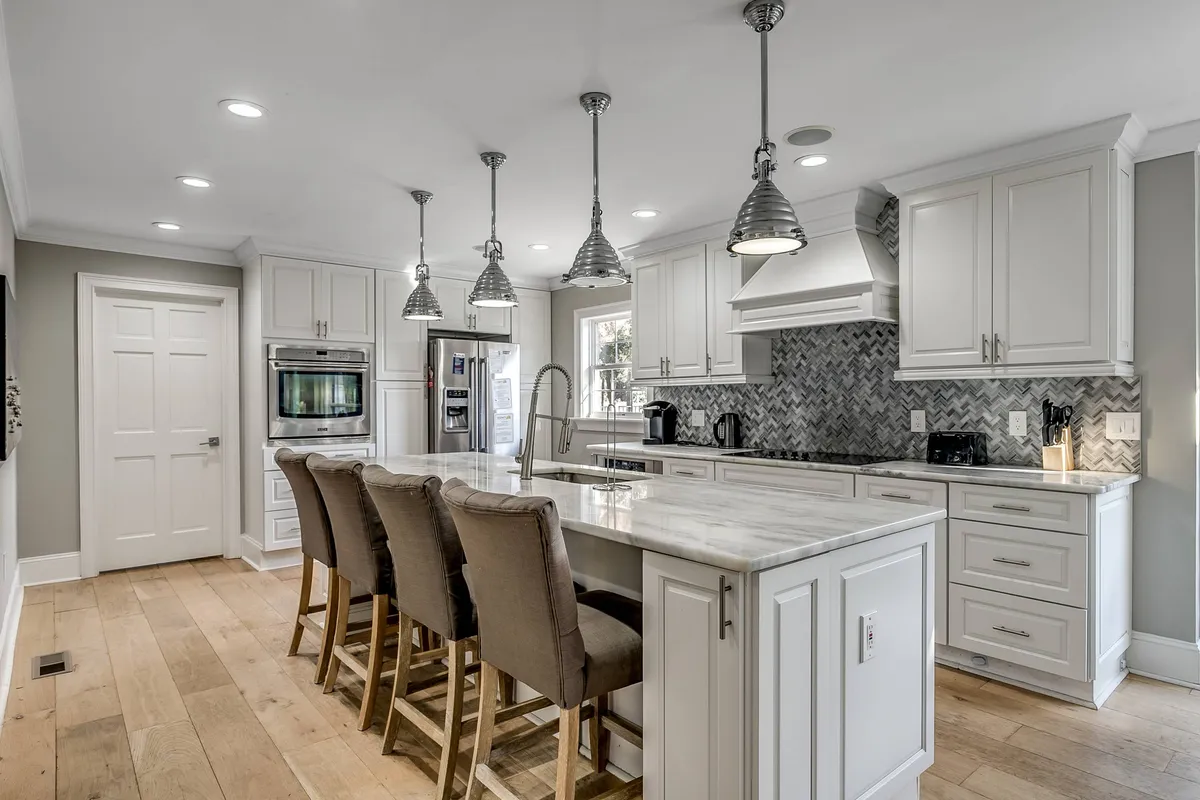 A spacious, upscale kitchen featuring an island counter with sleek bar stools, pendant lighting overhead, and a modern backsplash tile. Stainless steel appliances are visible, and the room exudes a sense of warmth and hospitality, perfect for both cooking and socializing.