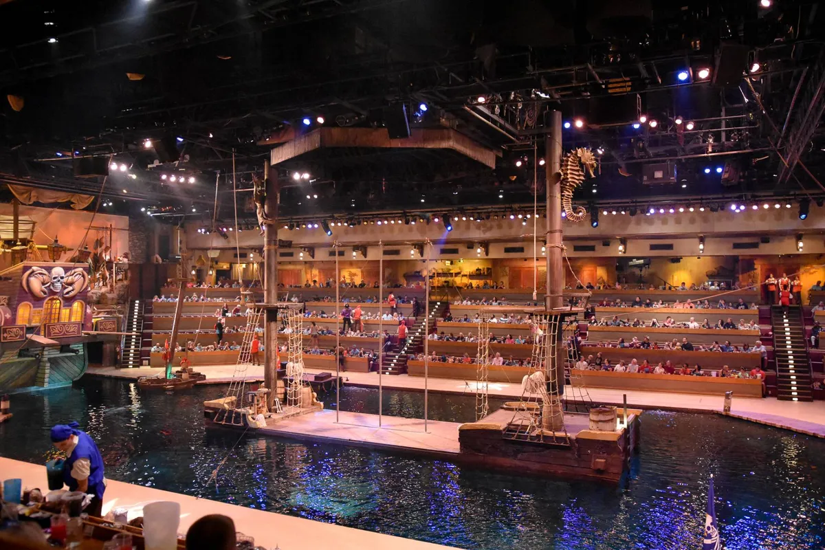 An interior shot of a vibrant dinner show scene, featuring a stage with a pirate ship theme, water feature, lively performers, and an engaged audience seated for dining. The festive lighting and theatrical setting create a sense of immersive entertainment and excitement.